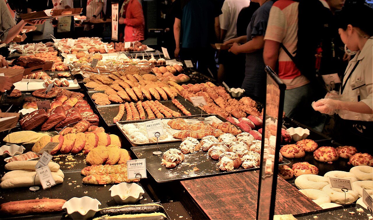Crowded bread display tables at Sungsimdang, customers with trays and staff restocking behind them