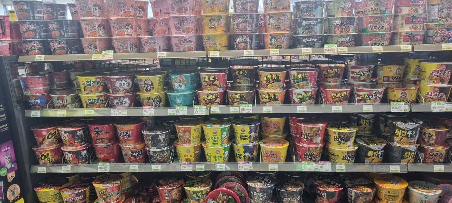 A wall of cup ramen in a Korean convenience store, with 1+1 and 2+1 price tags along the shelf edge