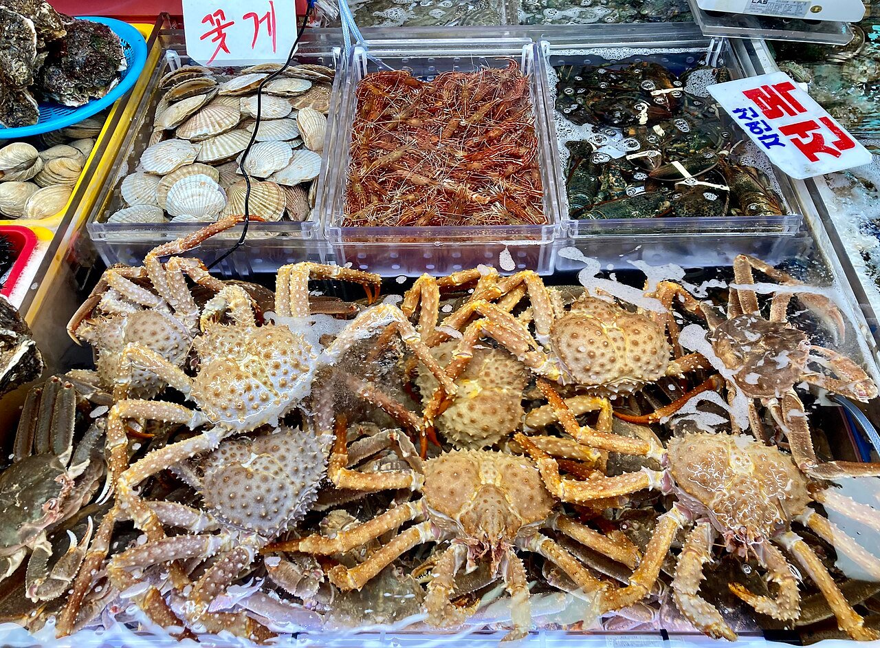 A market display of live crabs on ice with trays of scallops, clams, shrimp and lobsters above them and handwritten Korean price signs