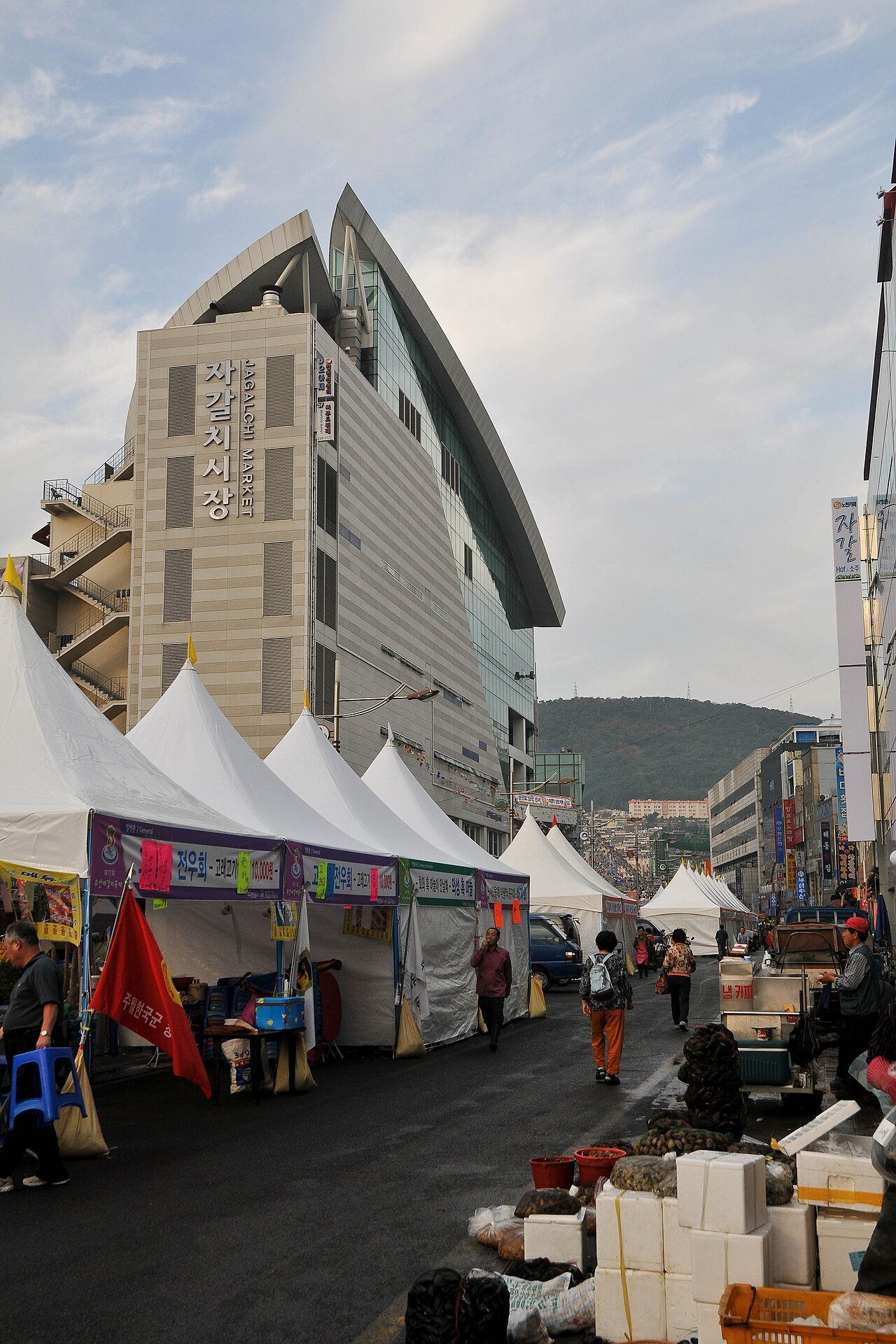 The Jagalchi Market building with its curved roof and Korean and English signage, white market tents and stacked polystyrene boxes along the street below it