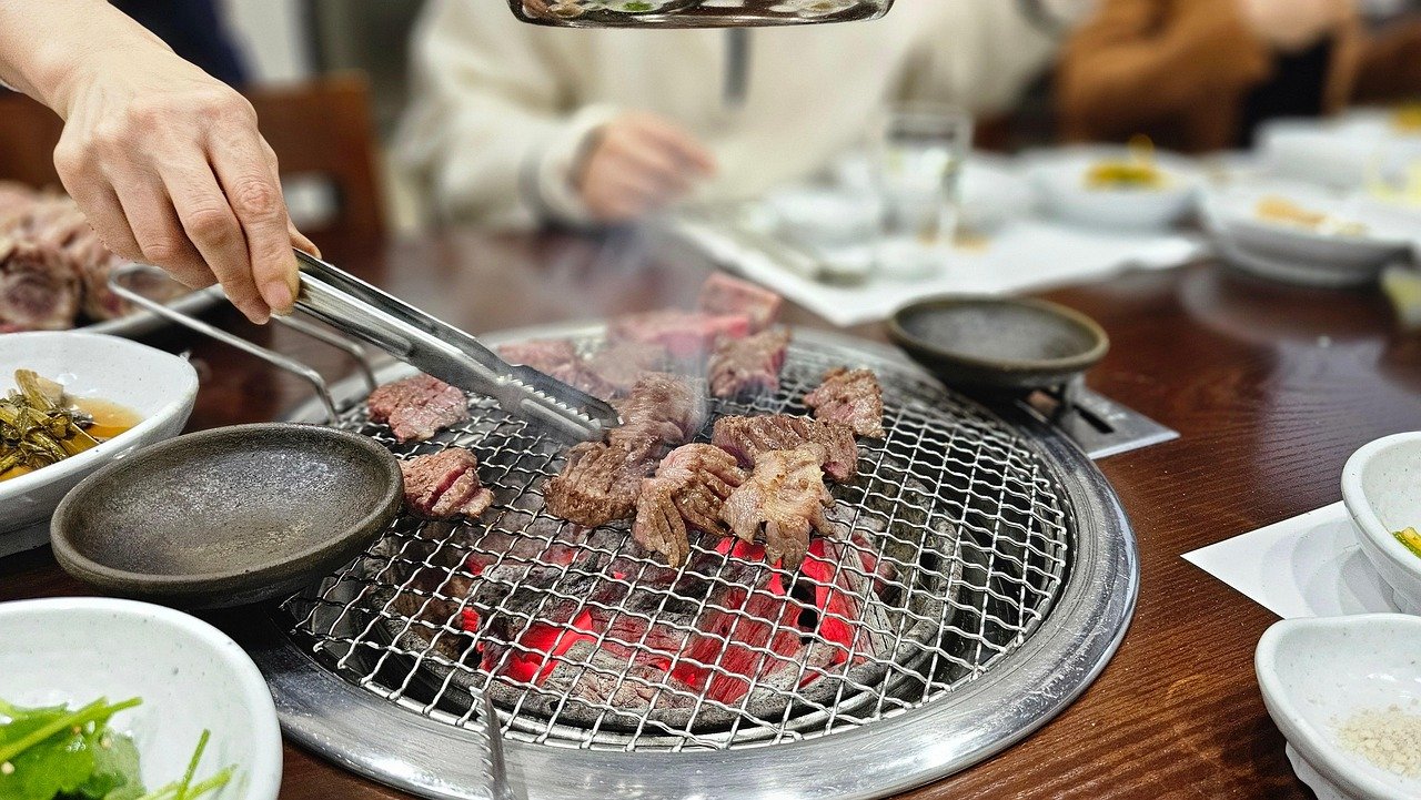A hand using metal tongs to turn beef on a charcoal grill at a Korean BBQ table, surrounded by small side dish bowls